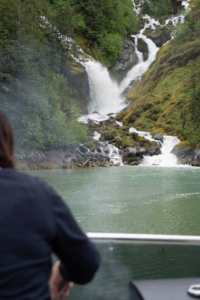 A guest on the deck of M/Y Dauntless watches a waterfall tumble down a mossy, forested cliff in LeConte Bay, Alaska.
