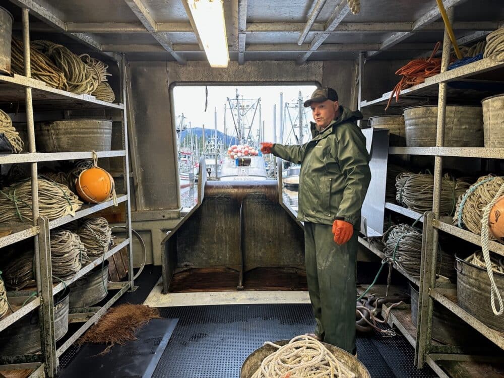 Captain Jim in rain gear aboard a fishing boat in Petersburg harbor, surrounded by coils of line and buoys, pointing out at the fleet. 