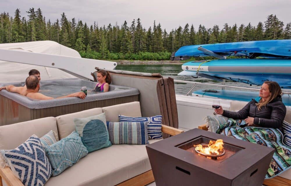 A family relaxes in the hot tub on the top deck of M/Y Dauntless while a guest sits by the fire pit, with kayaks racked behind them.